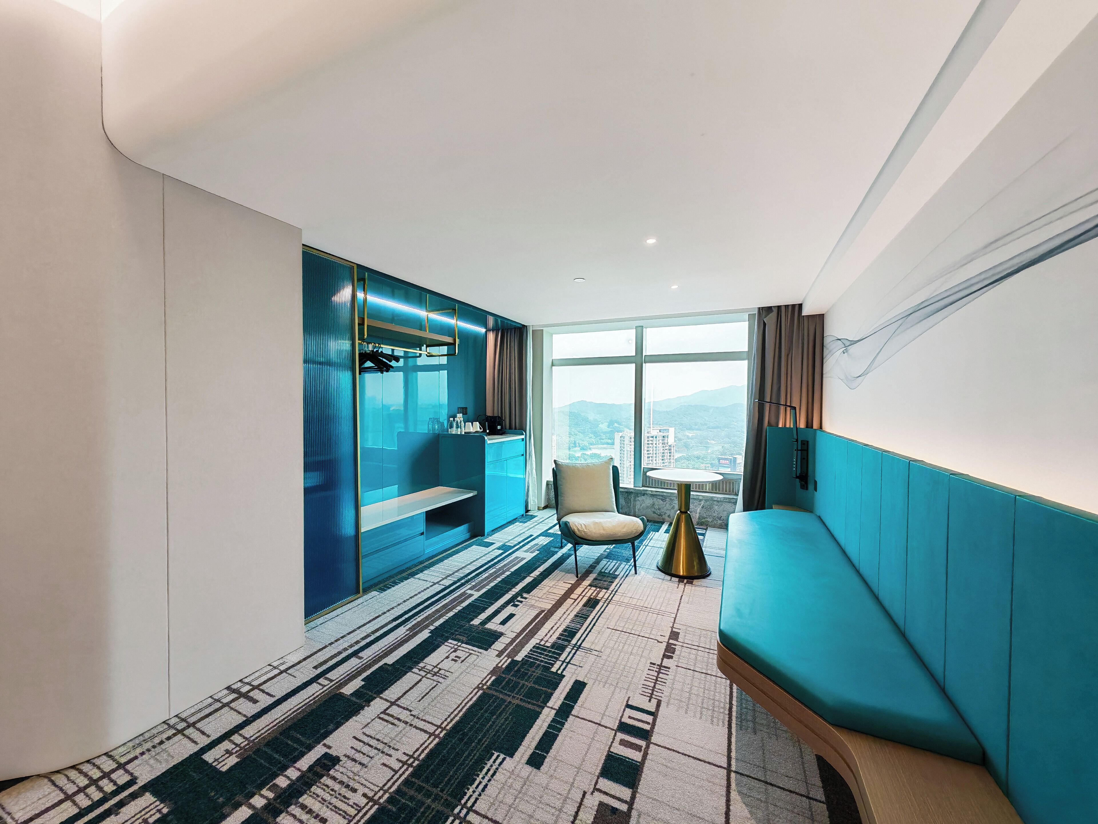 A modern hotel room with a large window, a seating area, a kitchenette, and a patterned carpet.