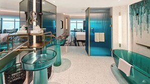 A hotel room with a glass partition, a bed, a bathroom with a blue wall, and a view of the city.