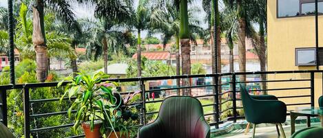 A covered outdoor seating area with a glass table and chairs, surrounded by potted plants and palm trees.