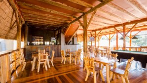 Uma área de bar com piso de madeira, mesas, cadeiras, mesa de bilhar e balcão com garrafas.