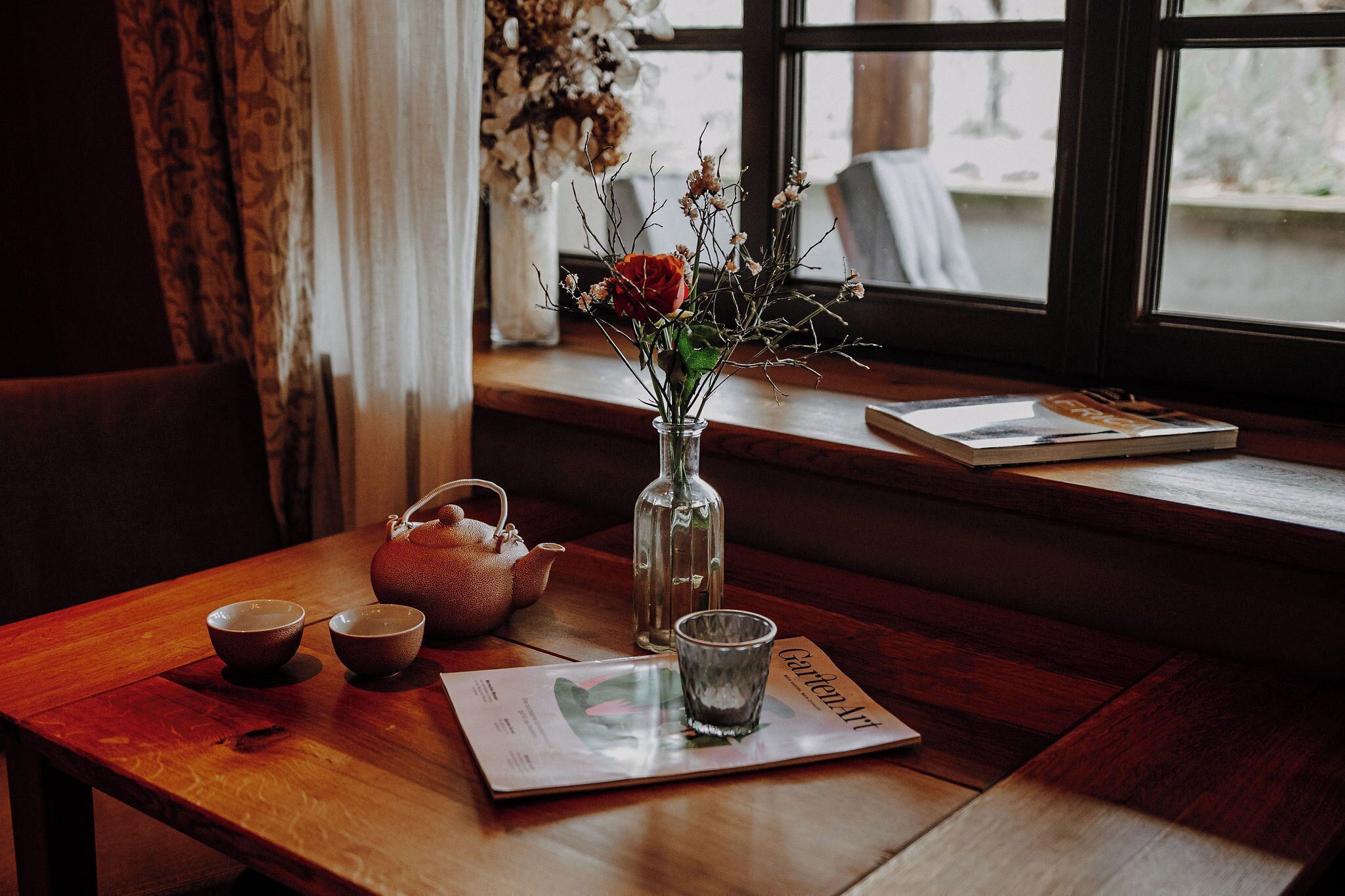 Una mesa de madera con un jarrón de flores, una tetera y dos tazas. Una revista titulada "Garden Art" está sobre la mesa.