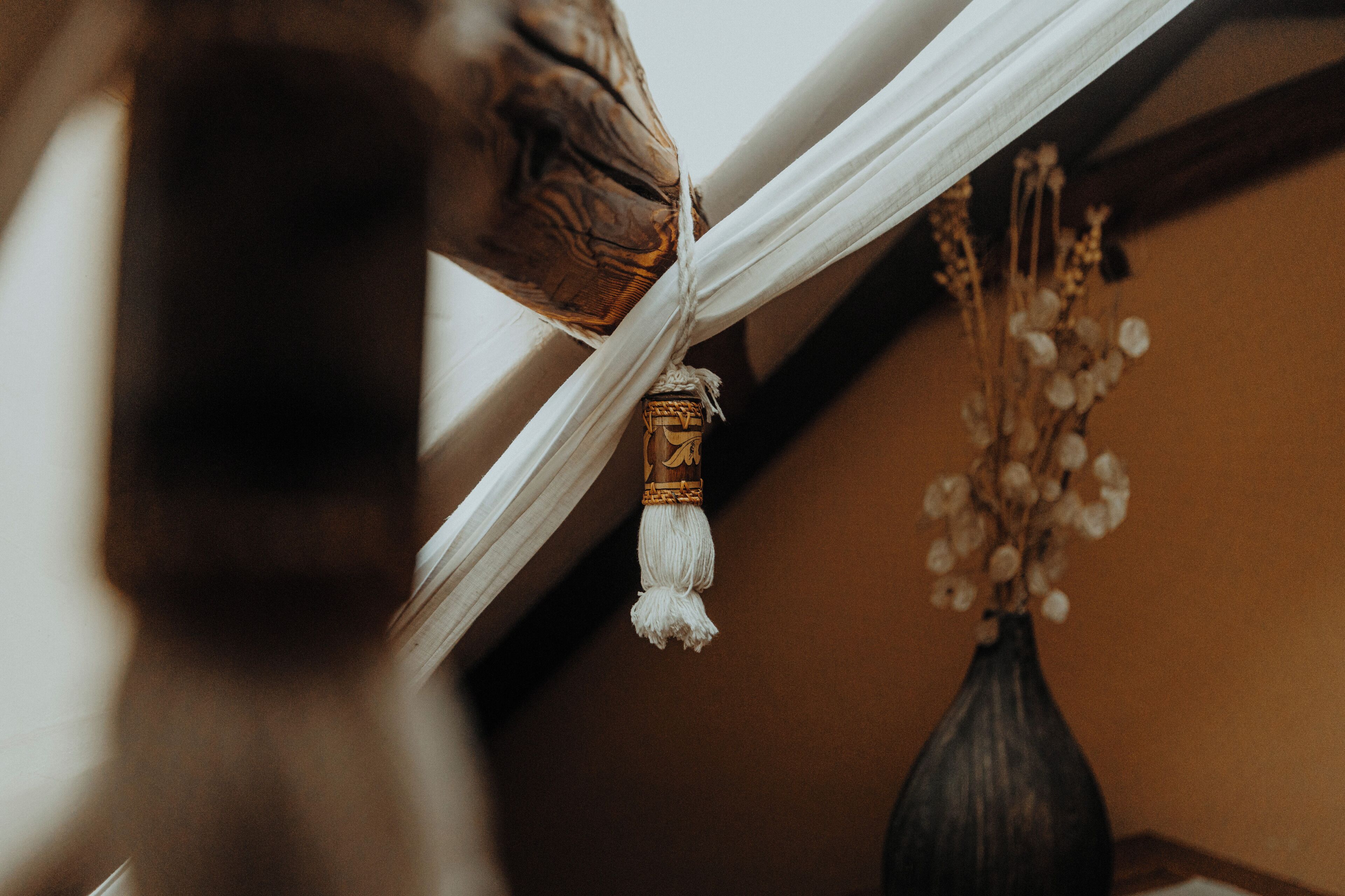 A close-up of a room corner with a white curtain, a decorative tassel, and a vase with dried flowers.