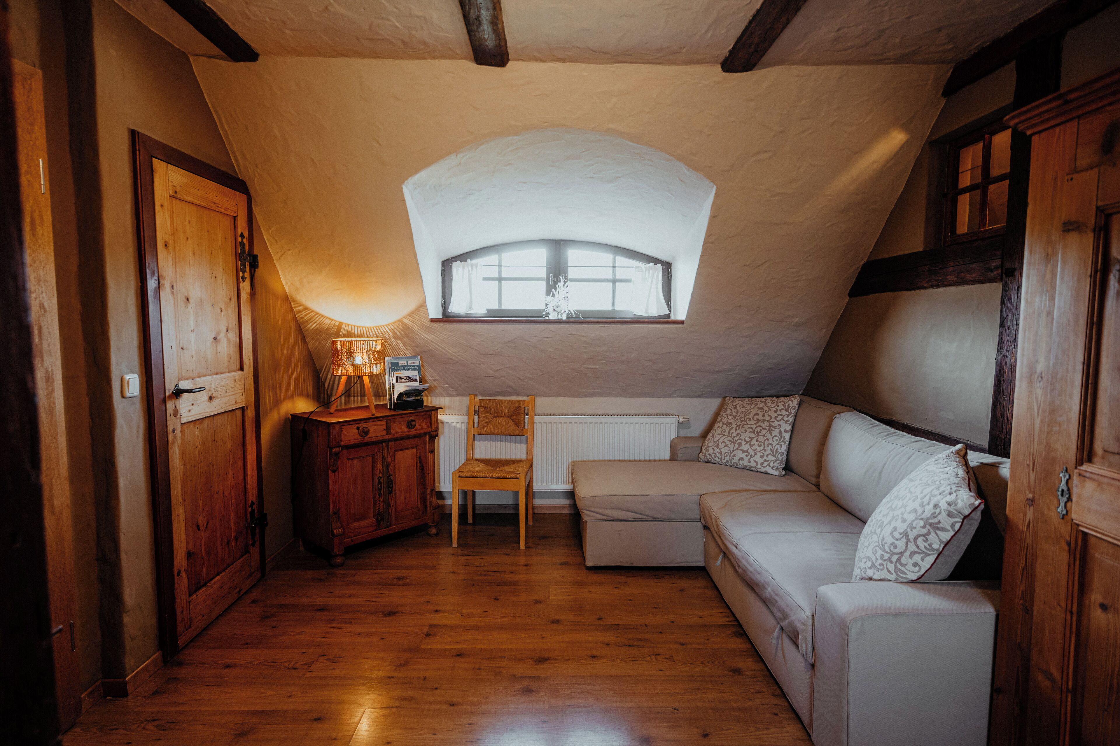 Un dormitorio acogedor en el ático, con techo inclinado, un armario de madera, una pequeña mesa con lámpara, un banco acolchado y una ventana con cortinas.