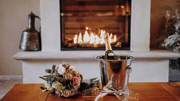 A wooden table with a champagne bottle in a metal bucket, a bouquet of flowers, and a fireplace in the background.