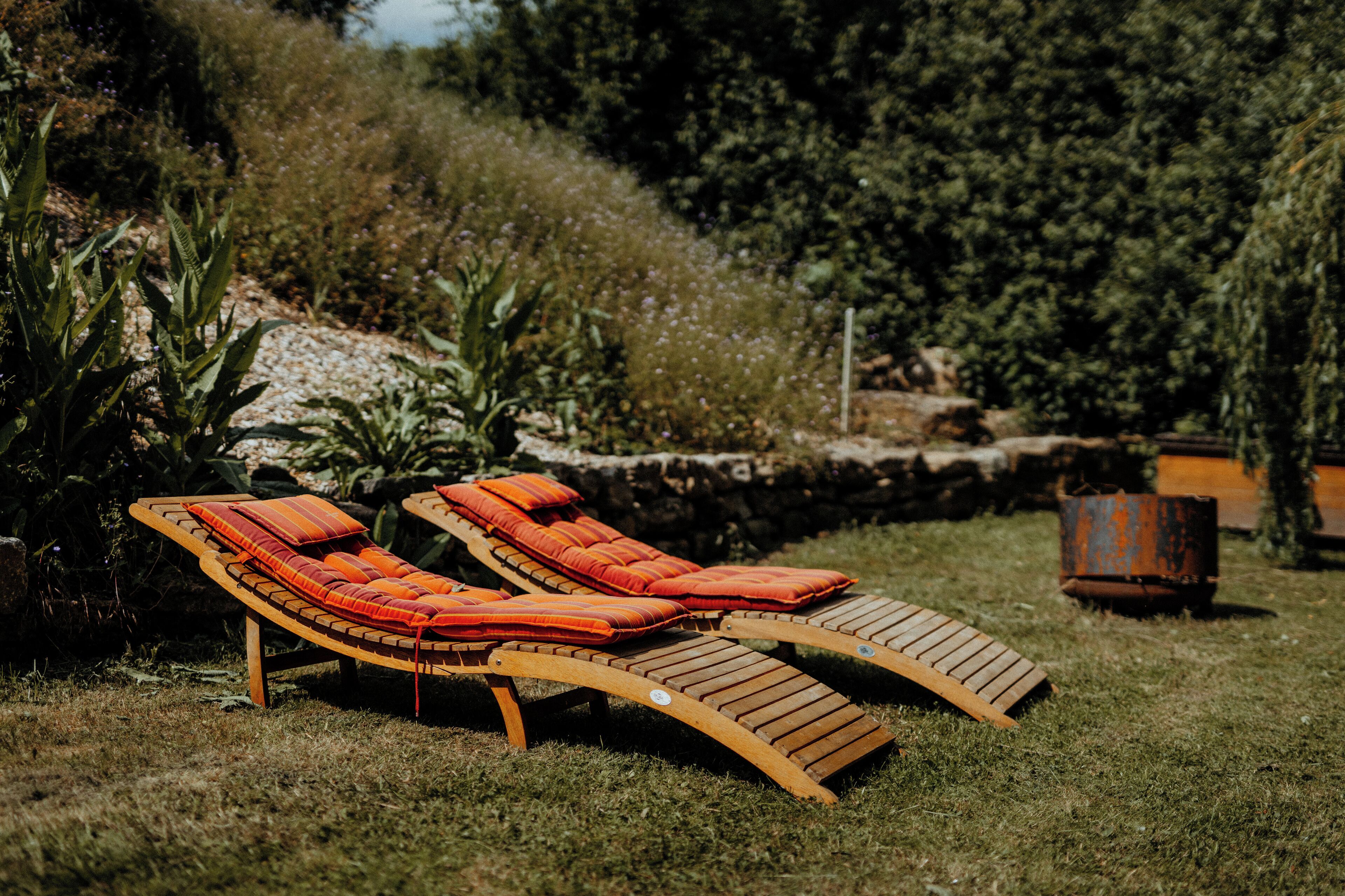 Zwei Holz-Loungestühle mit orangefarbenen Kissen in einer grünen Außenanlage mit einer Steinmauer und Bepflanzung im Hintergrund.