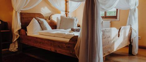 A wooden bed with white bedding and a canopy, a wooden headboard, and a window with a view of greenery.