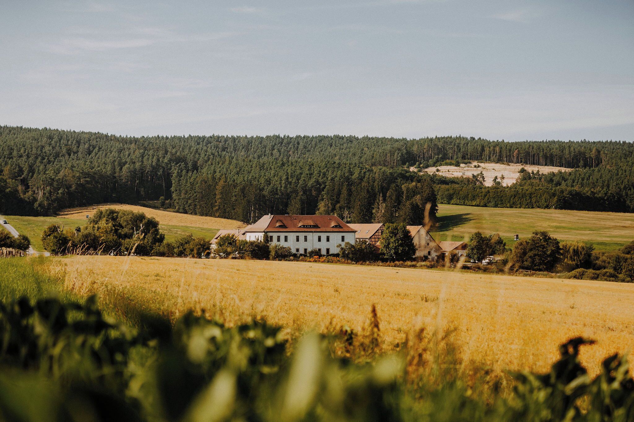 Ett lantligt landskap med en vit byggnad omgiven av åkrar och en skogbeklädd kulle.