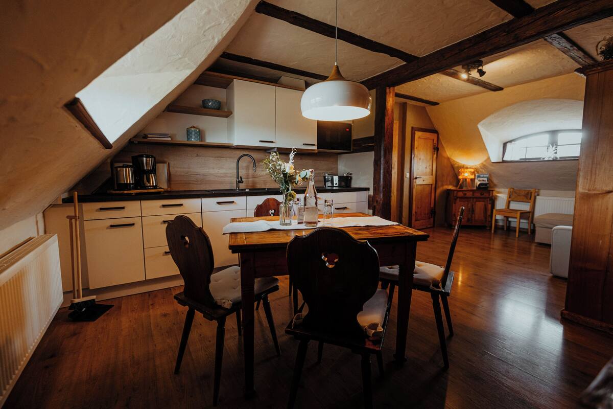 A cozy kitchen with wooden cabinets, a dining table, and chairs. There is a ceiling fan and a window providing natural light.