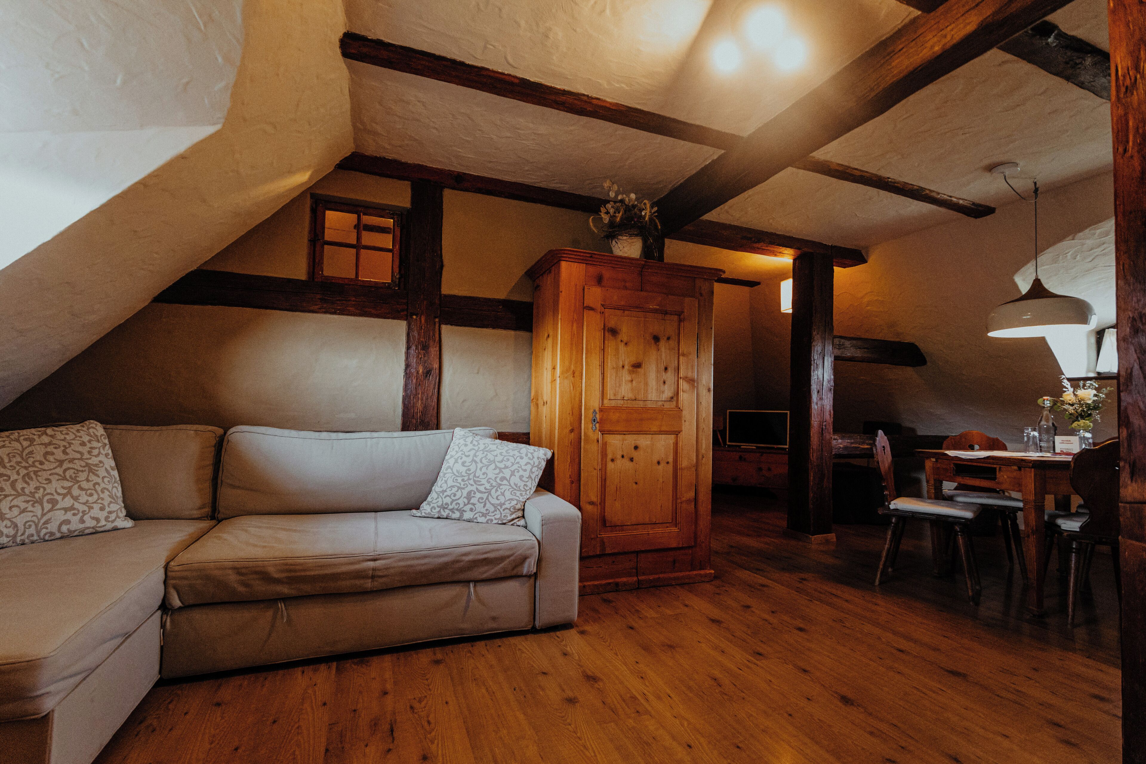 A cozy attic room with a wooden ceiling, a sofa, a cabinet, and a dining table with chairs.