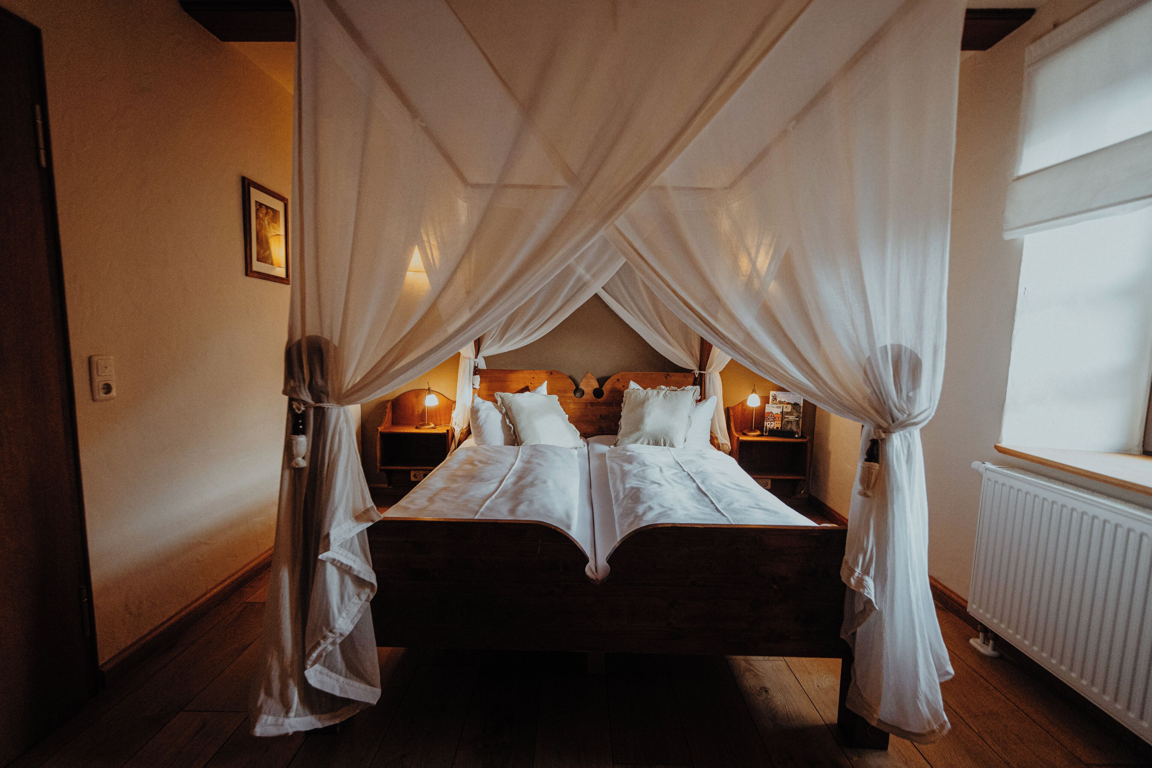 A bedroom with a wooden bed, white bedding, and a canopy. There are bedside lamps and a framed picture on the wall.