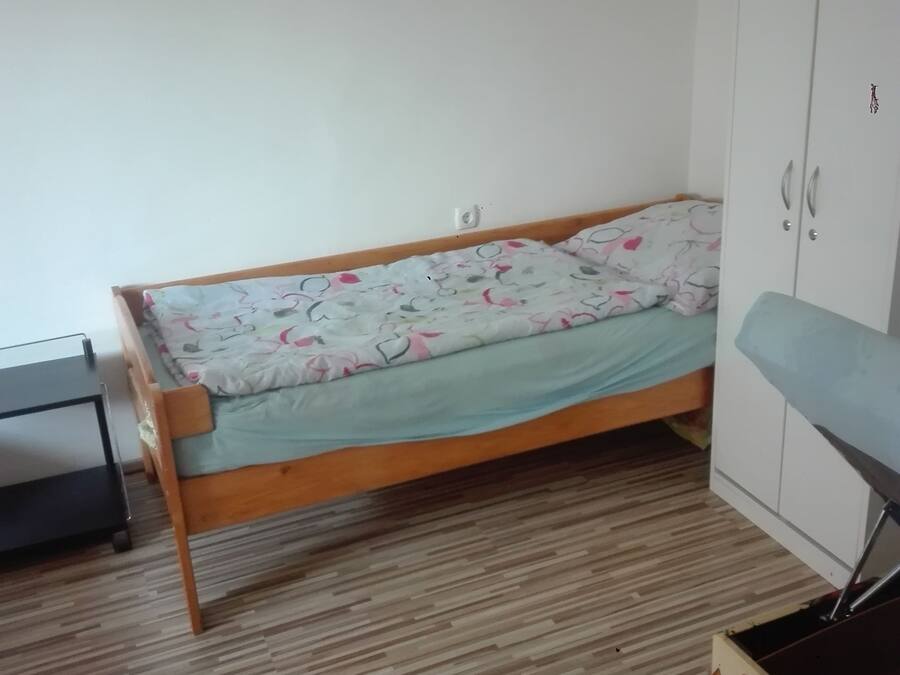 A single bed with a floral bedspread, a wooden floor, a wardrobe, and a small table.