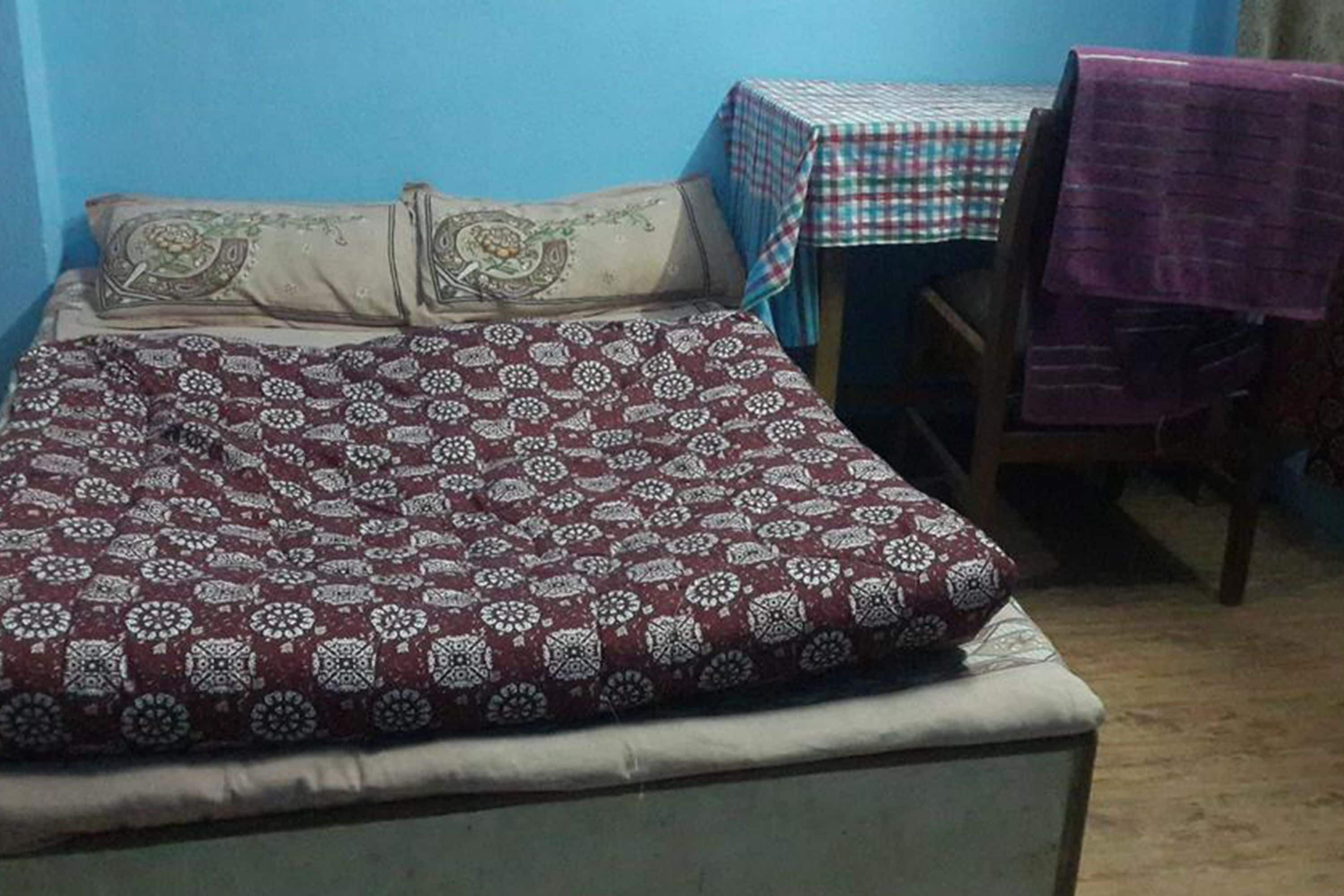 Un lit recouvert d’une couvre-lit à motifs, une table avec une nappe à carreaux et une chaise recouverte d’un tissu violet.