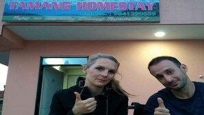 Two people sitting at a table with a sign above them that reads "TAMANG HOMESTAY" and a phone number. - Tamang Homestay (Dhulikhel)