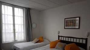 Une chambre d’hôtel avec deux lits simples, une grande fenêtre avec des rideaux légers, un tableau accroché au mur et un miroir rond.