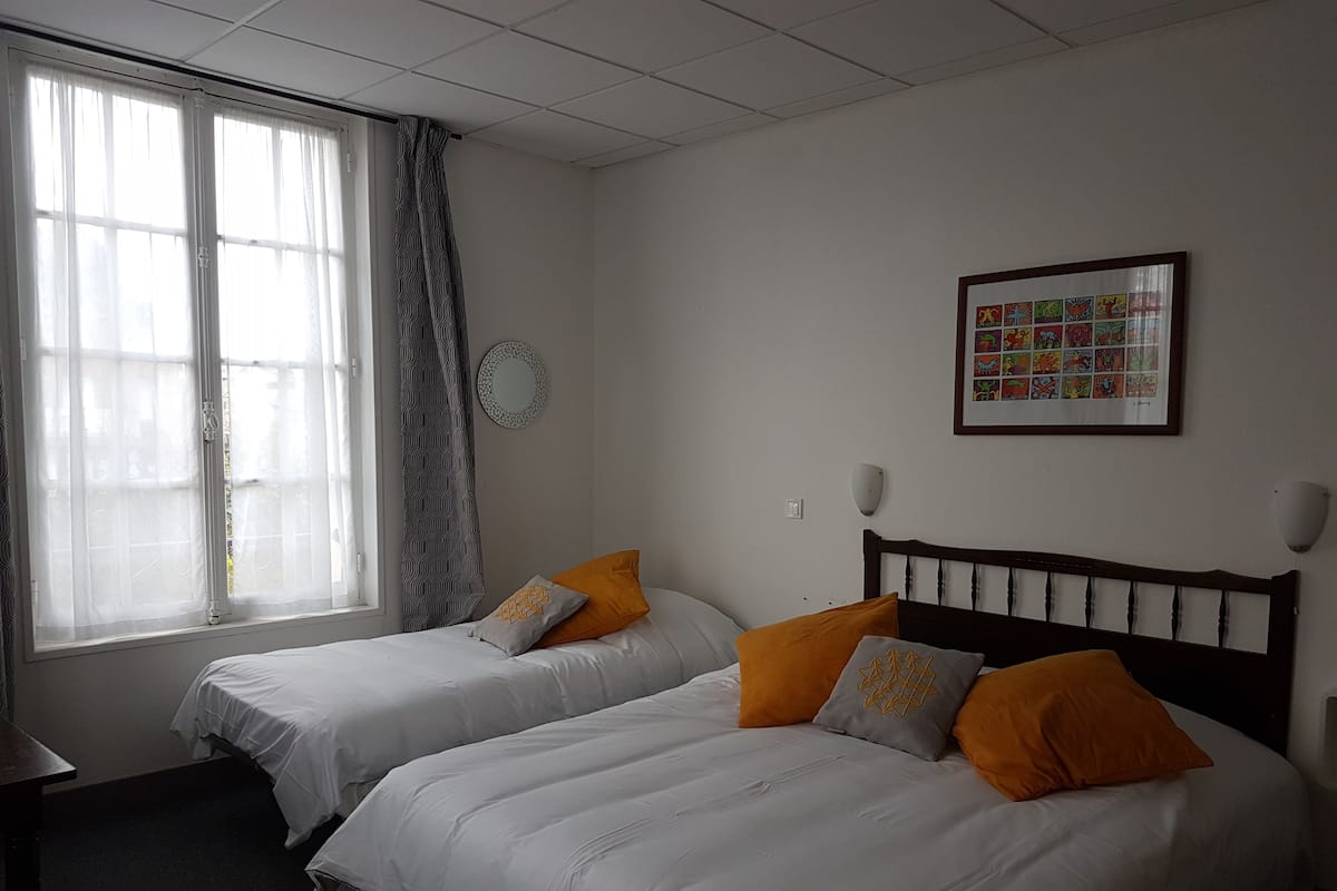 A hotel room with two single beds, a large window with sheer curtains, a wall-mounted picture, and a round mirror.