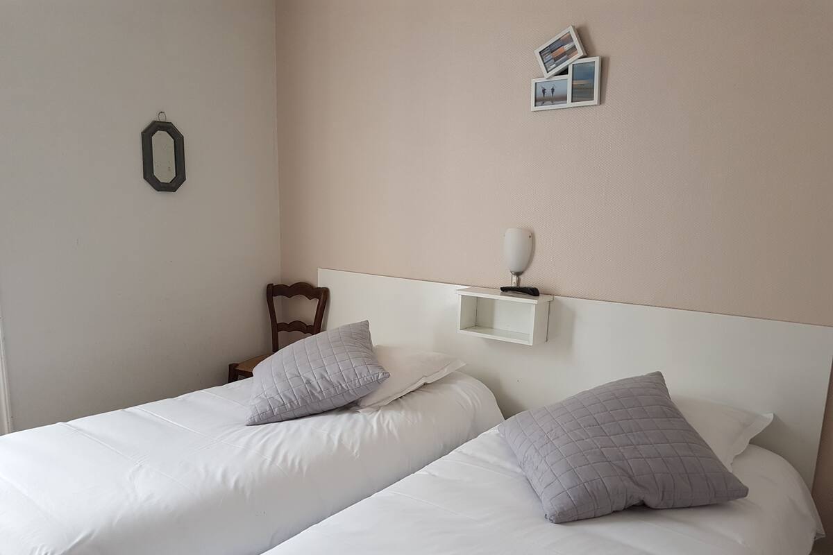 A hotel room with two single beds, each with a white mattress and a grey quilted pillow. There is a small bedside table with a lamp on it, and a wall-mounted shelf above the beds.