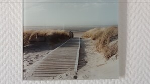 Une photographie encadrée représentant une promenade en bois menant à une plage bordée de dunes.