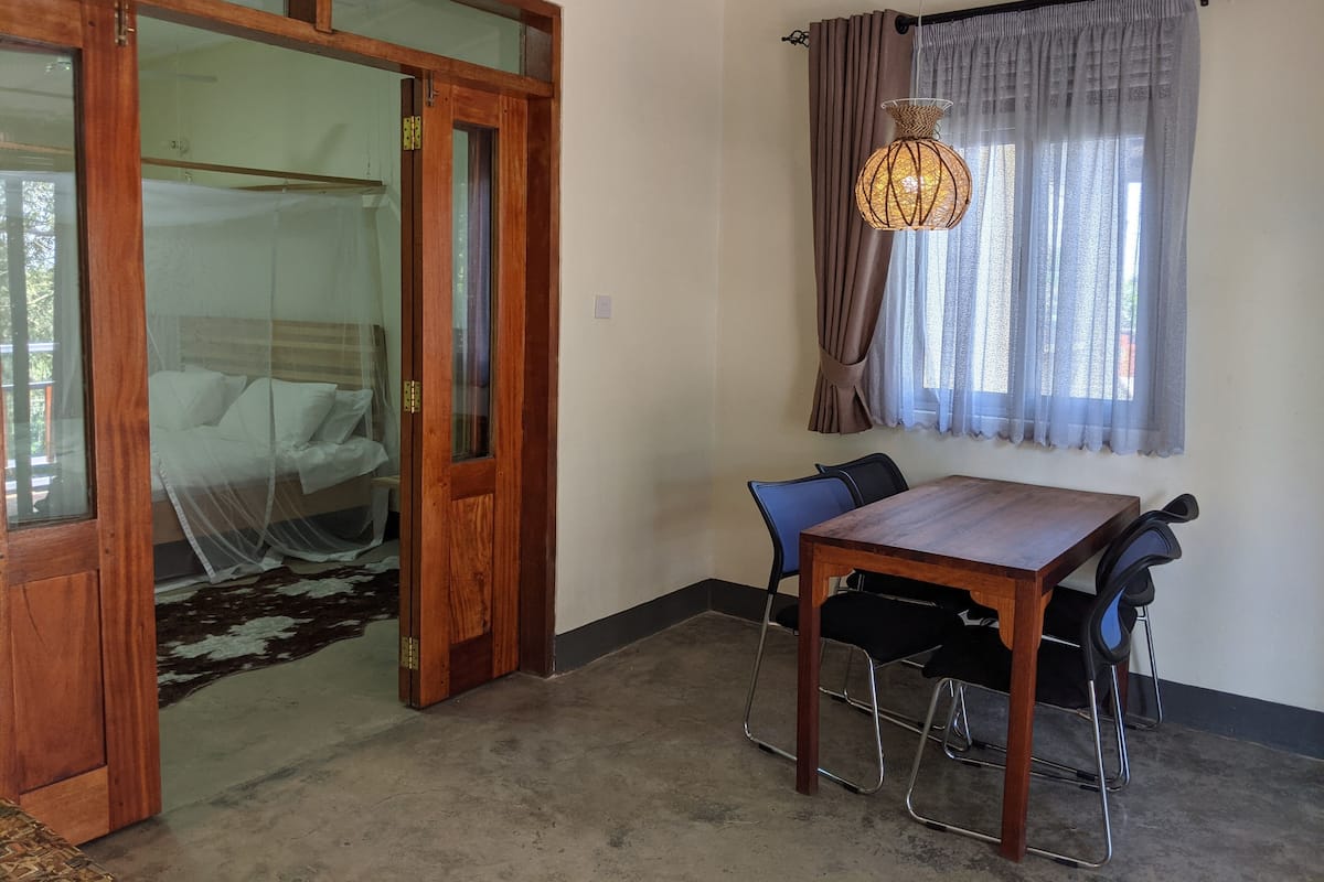 A room with a wooden door, a bed with white linens, a dining table with chairs, and a hanging light fixture.