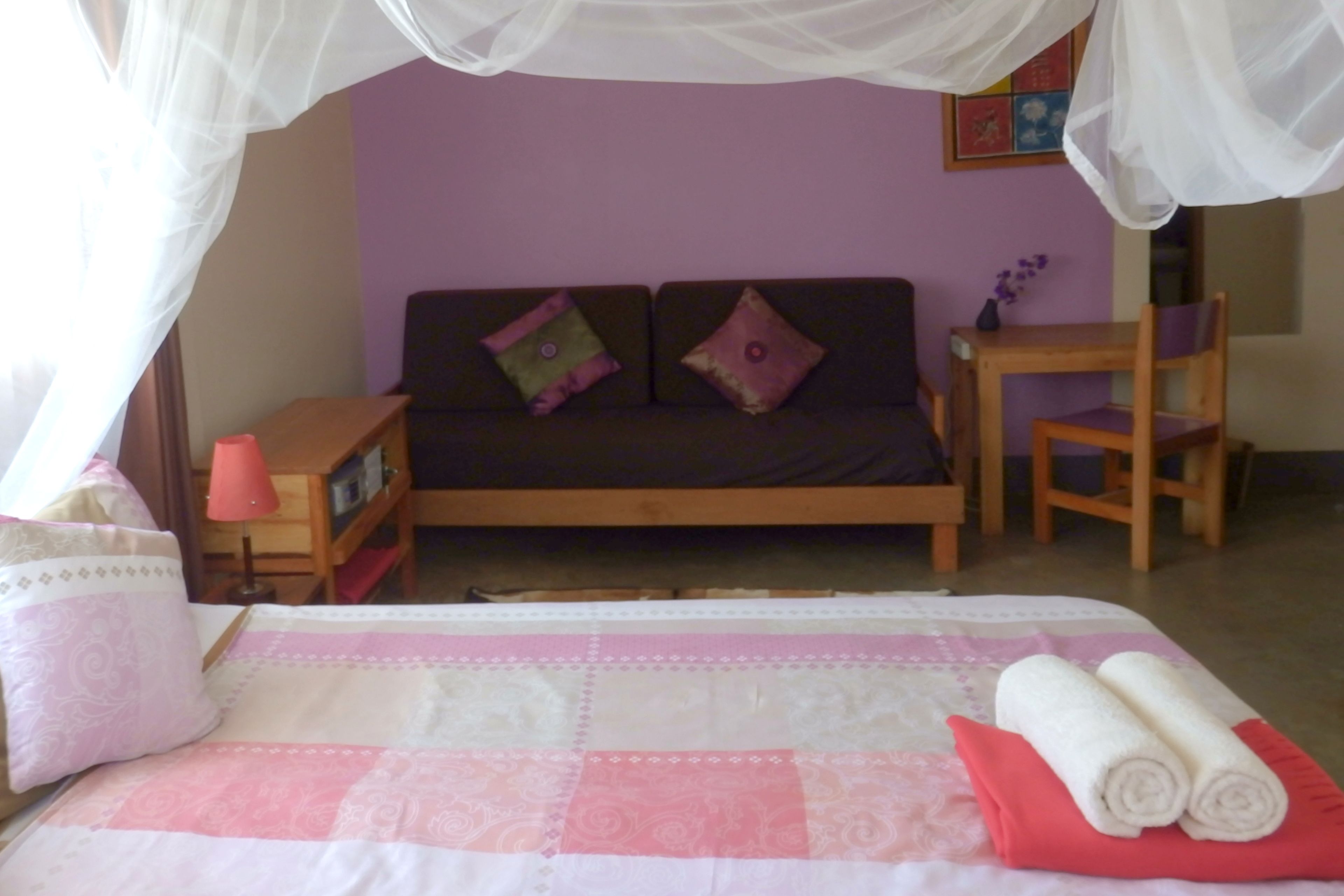 A bedroom with a bed, a nightstand, a wooden chair, a small table, and a mosquito net over the bed.