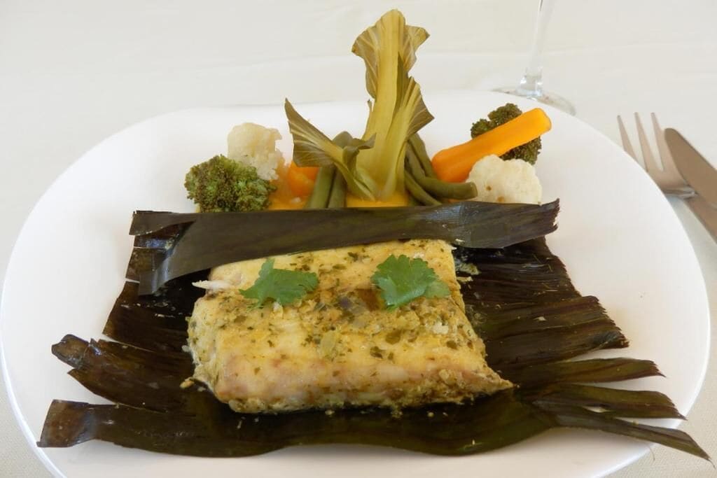 A plate with a piece of fish on banana leaves, accompanied by vegetables and a glass of water.