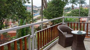 Varanda com cadeira de vime e mesa de vidro, com vista para área residencial com árvores e edifícios.