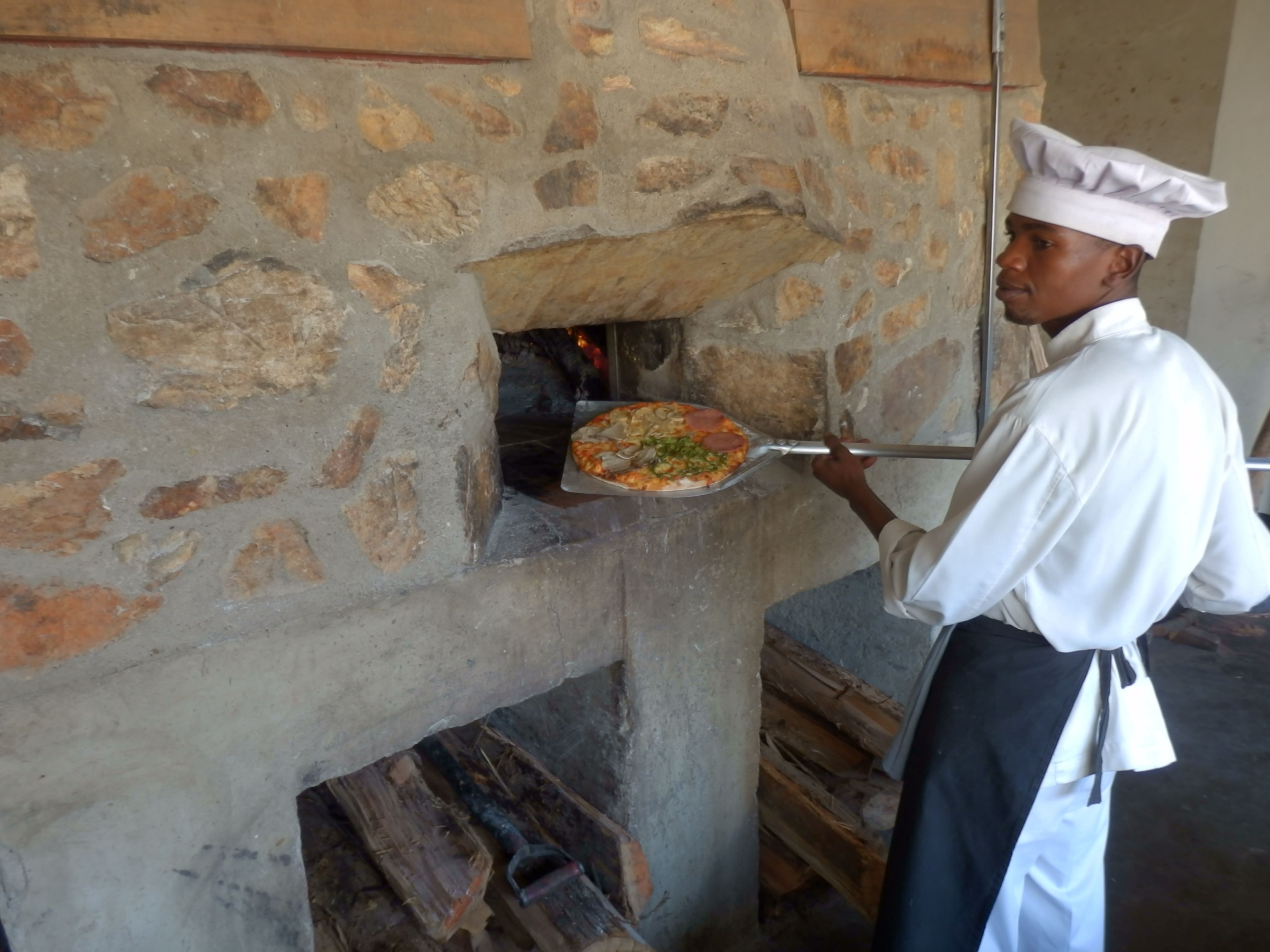 A chef in a white uniform and hat, using a pizza peel to place a pizza into a stone oven.