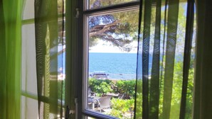 Vista per una finestra amb cortines verdes, sobre una platja amb mar blau turquesa, un litoral rocosos i vegetació.