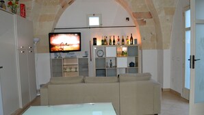 A living room with a glass-top coffee table, beige sofas, a TV mounted on the wall, and a wine rack with bottles. - Le Terrazze B&B (Manduria)