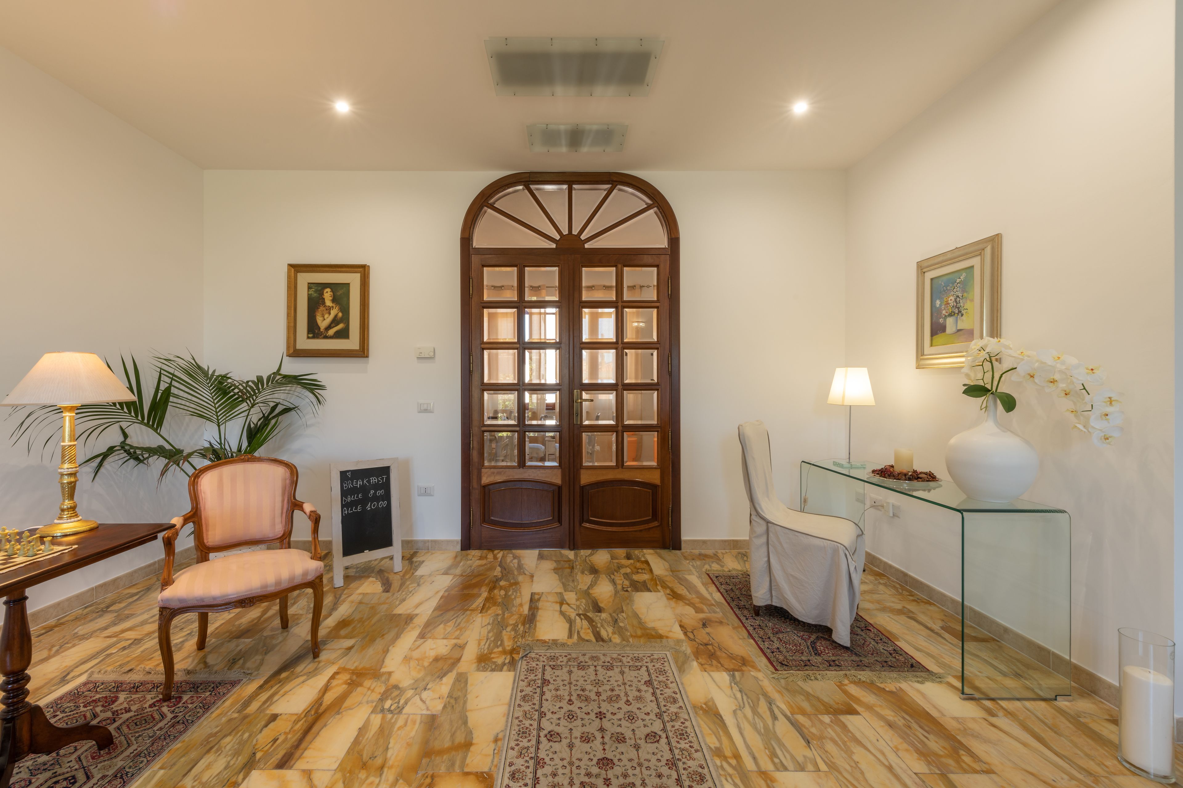 A hotel lobby with a wooden door, a table with a lamp, a chair, a glass table with a vase, and a rug.