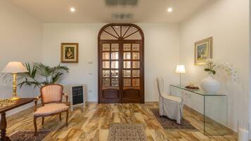 A hotel lobby with a wooden door, a table with a lamp, a chair, a glass table with a vase, and a rug.