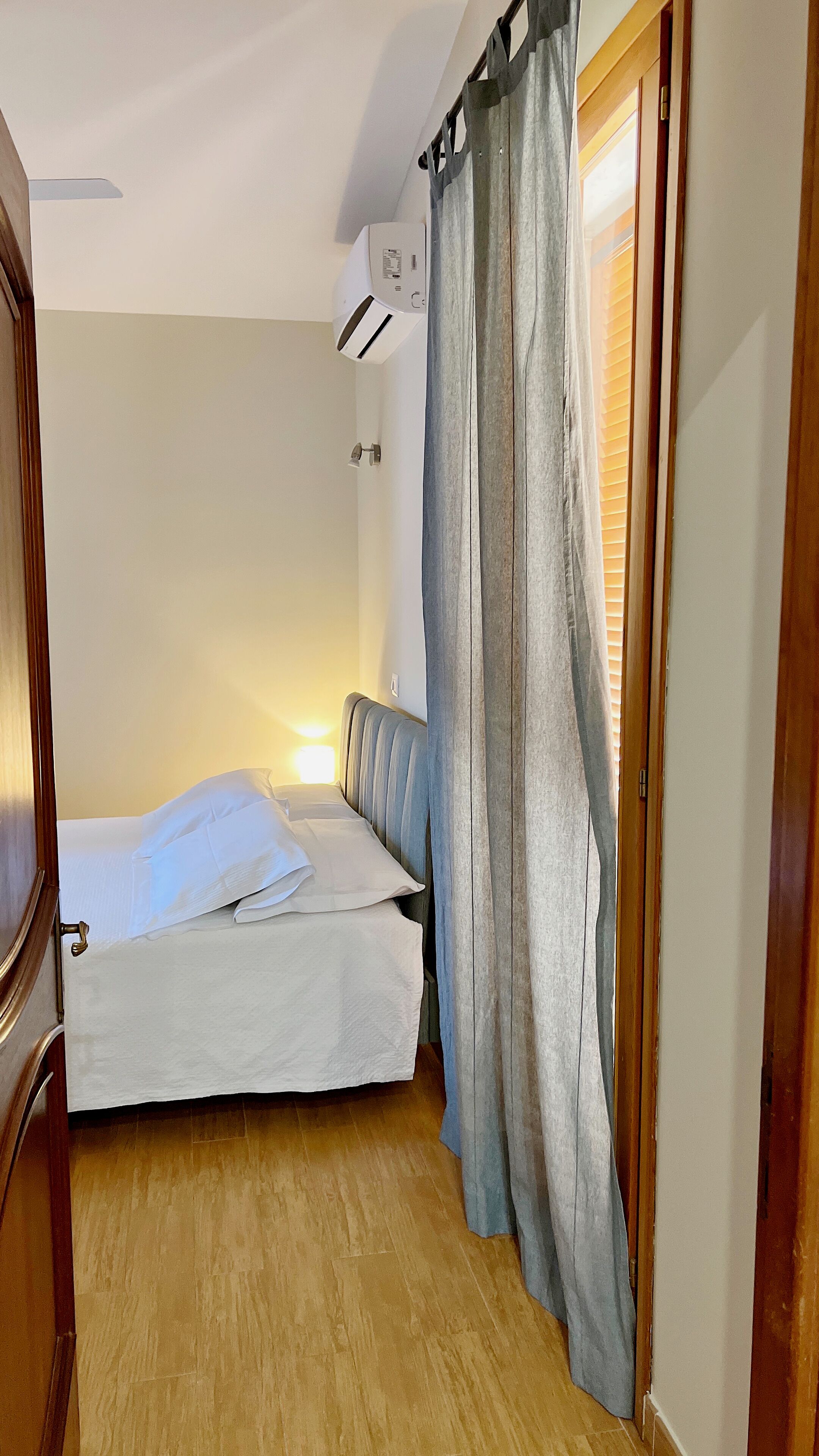 Ein kleines, ordentlich bezogenes Bett mit weißen Leinen, Holzboden und einem Fenster mit Jalousien.