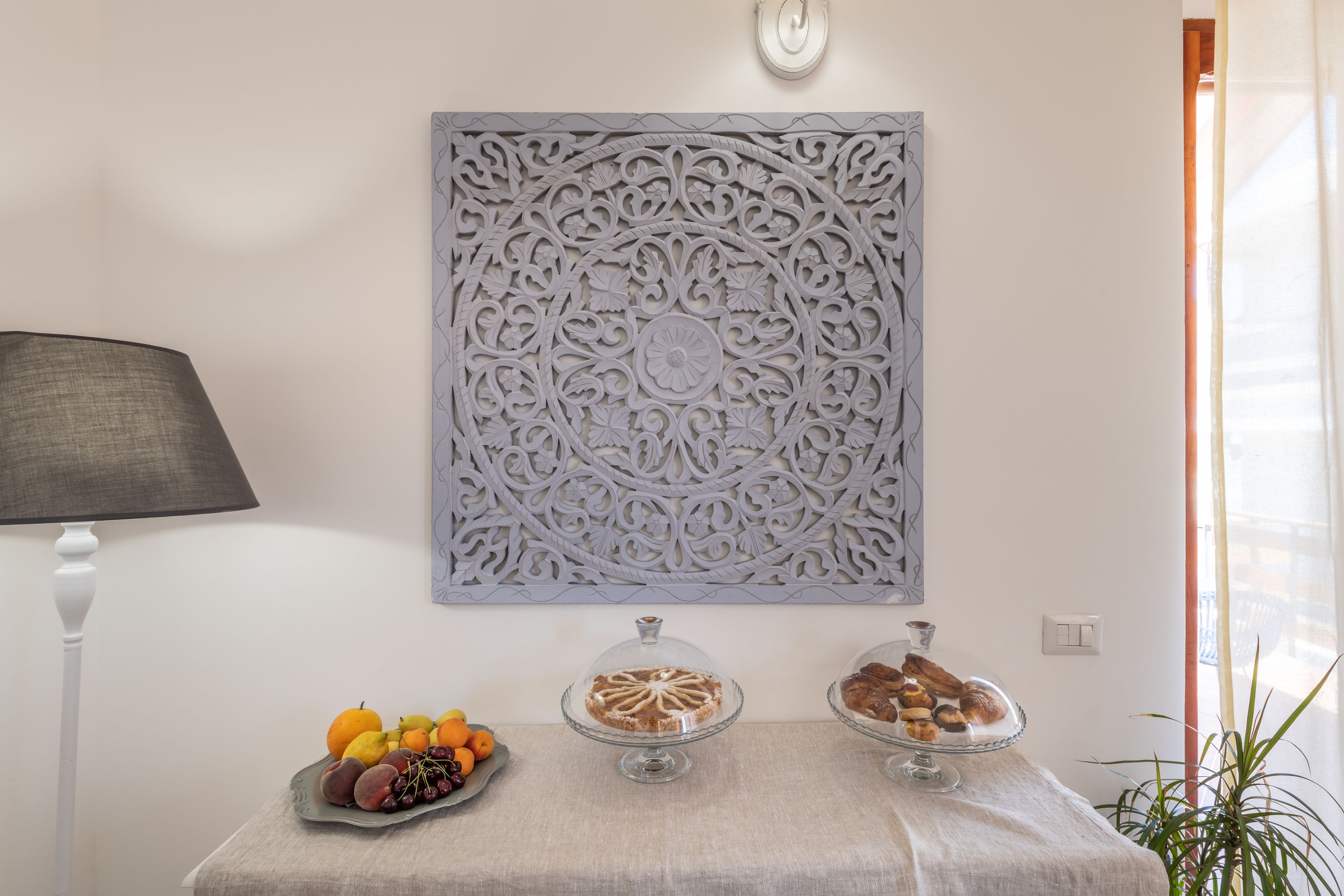 A table with fruit and pastries, a decorative wall panel, and a lamp.