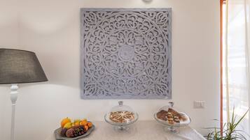 A table with fruit and pastries, a decorative wall panel, and a lamp.