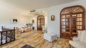 A spacious living area with a marble floor, a wooden staircase, a white sofa, a coffee table, and a large wooden door with glass panels. - Villa Iole (Carloforte)