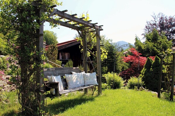 A wooden pergola with a swing bench, surrounded by lush greenery and flowering plants. - Pension Sonnenzeit (Reit im Winkl)