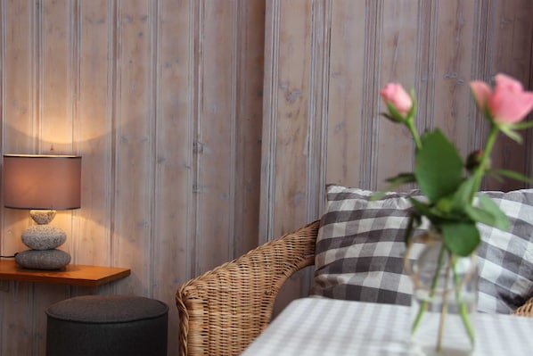 Una habitación acogedora con una silla de mimbre, un cojín de cuadros, una mesita con una lámpara y un jarrón con flores.