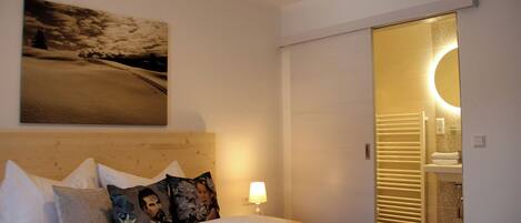A hotel room with a bed, pillows, a wall-mounted painting, a bedside lamp, and a bathroom visible through an open door.