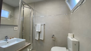Un bany modern amb un plat de dutxa de vidre, un lavabo blanc amb taulell de fusta, un vàter i una televisió penjada.