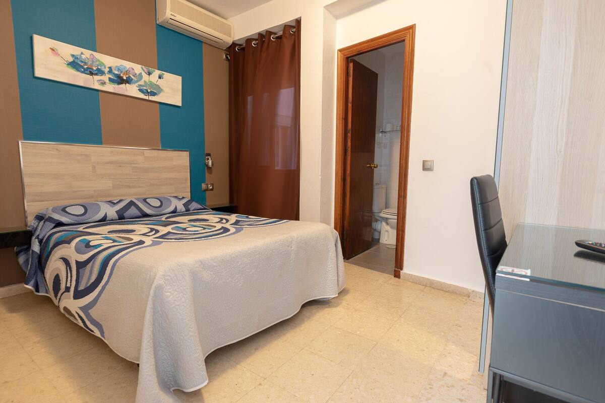 Habitación de hotel con cama, escritorio, silla, puerta de baño y un cuadro en la pared.