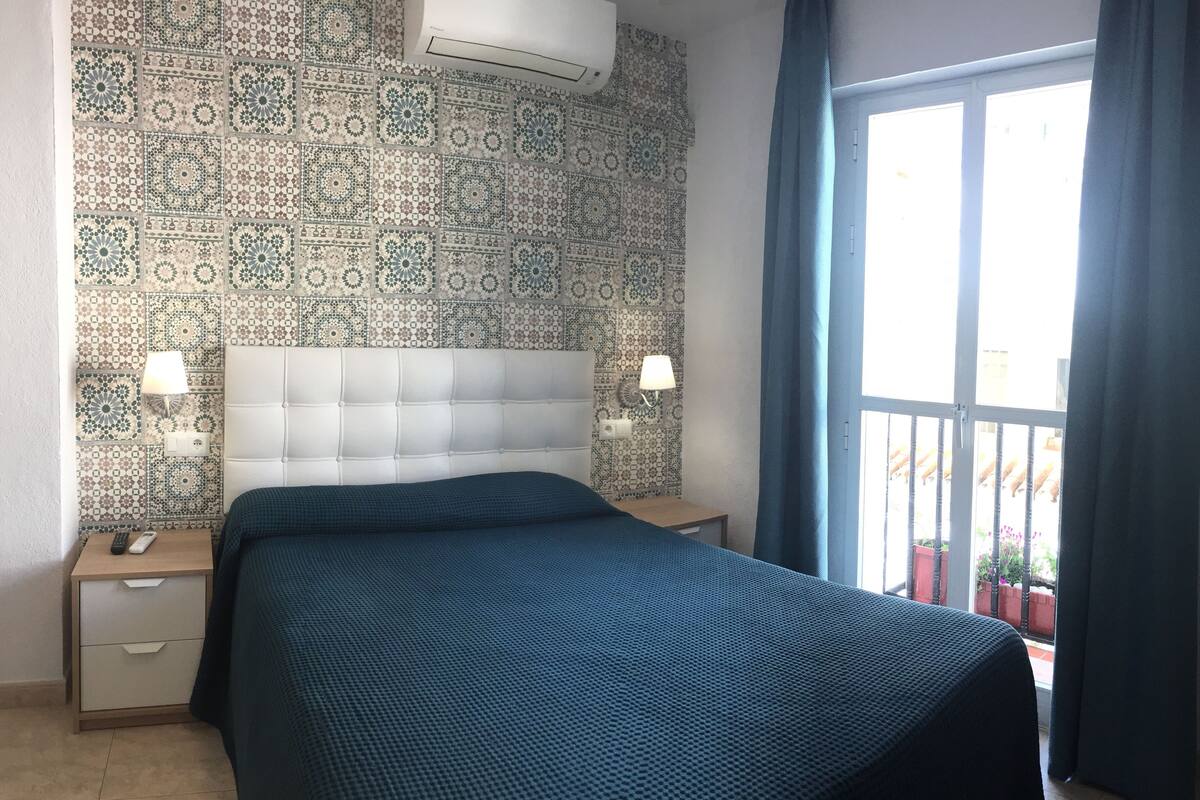 Habitación de hotel con pared estampada, una cama grande, dos mesitas de noche con lámparas y vistas al exterior a través de una puerta corredera.