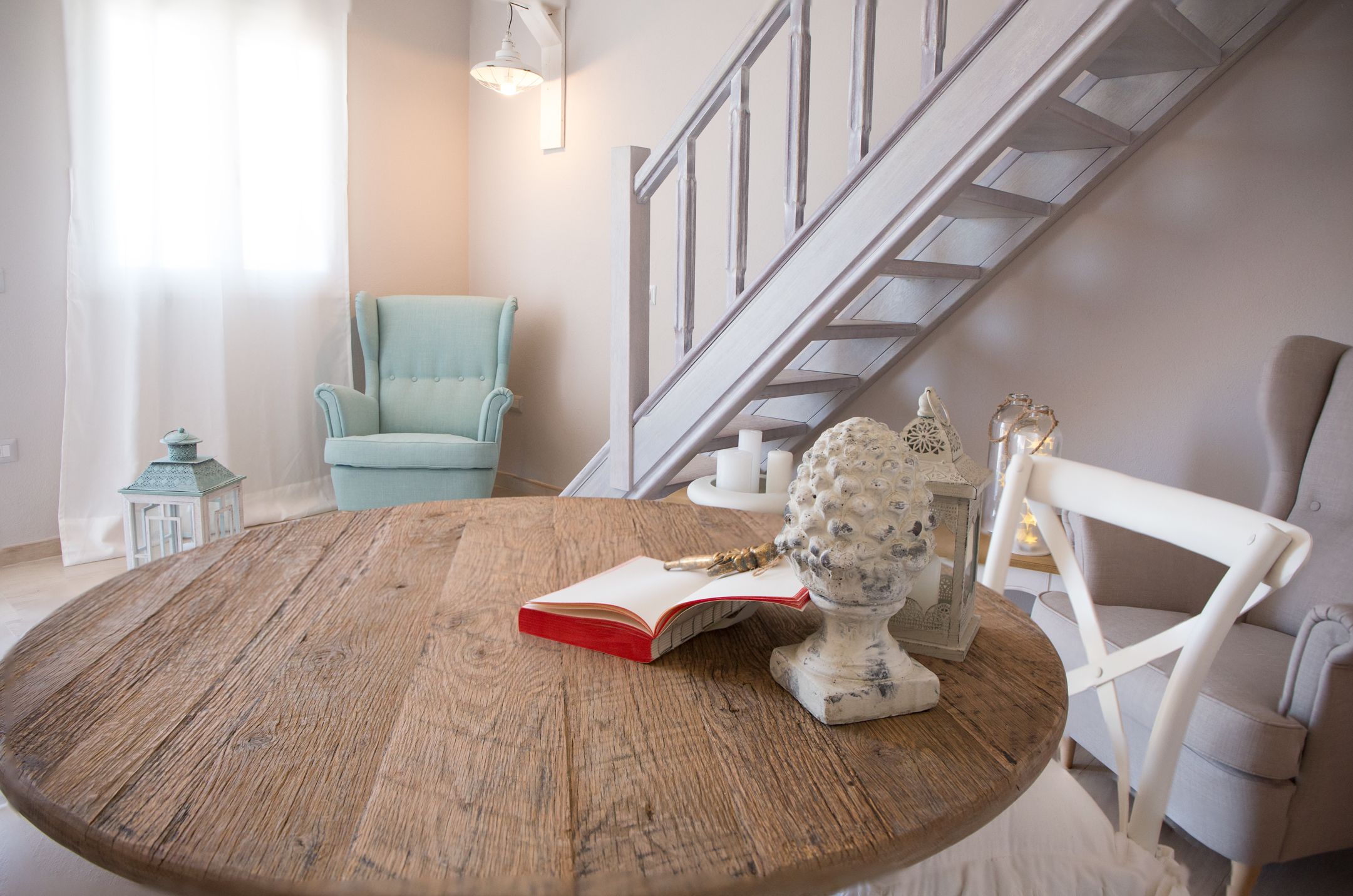 A round wooden table with a book and a decorative item. A light blue armchair and a white chair are visible. A staircase with a wooden railing is in the background.