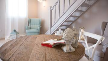 A round wooden table with a book and a decorative item. A light blue armchair and a white chair are visible. A staircase with a wooden railing is in the background.