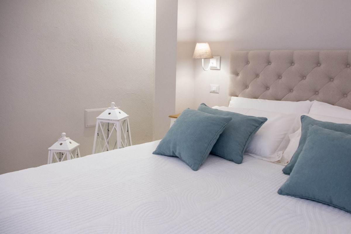 A neatly made bed with white linens and blue pillows, a tufted headboard, a wall-mounted lamp, and a white lantern on a bedside table.