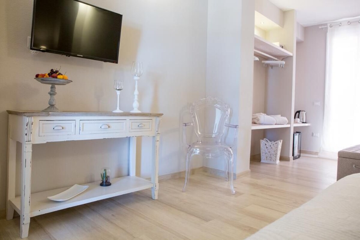 A hotel room with a white desk, a clear chair, a television mounted on the wall, and a closet with shelves.