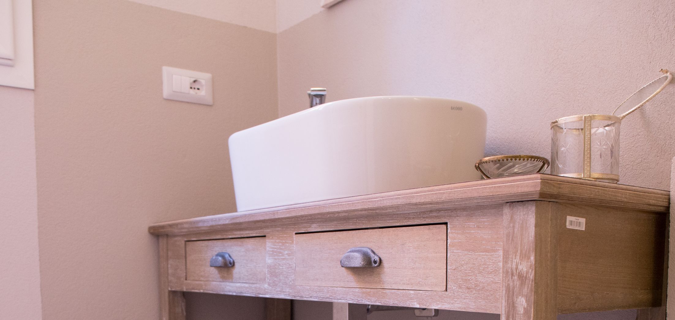 Un tocador de baño con un lavamanos blanco, un armario de madera y un recipiente de vidrio encima.