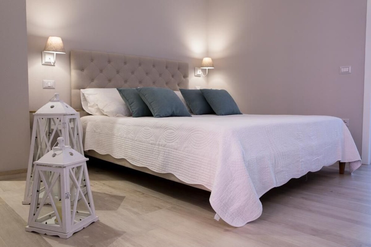 A neatly made bed with a white duvet and blue pillows, a white upholstered headboard, and two wall-mounted lamps.