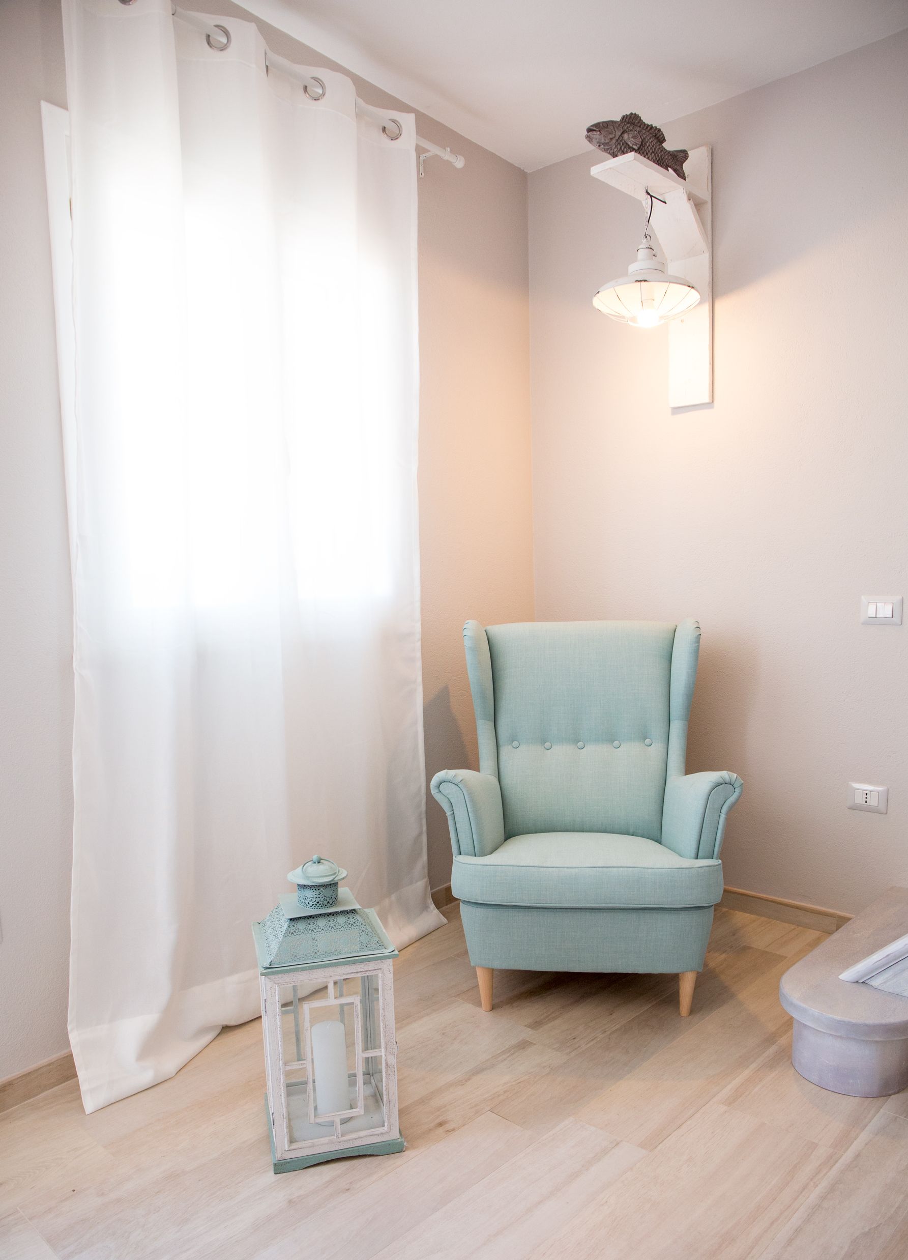 A room with a light blue armchair, a white curtain, a wall-mounted lamp, and a small white lantern on the floor.