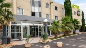 Een modern hotel met een glazen ingang, palmbomen en een parkeerplaats.