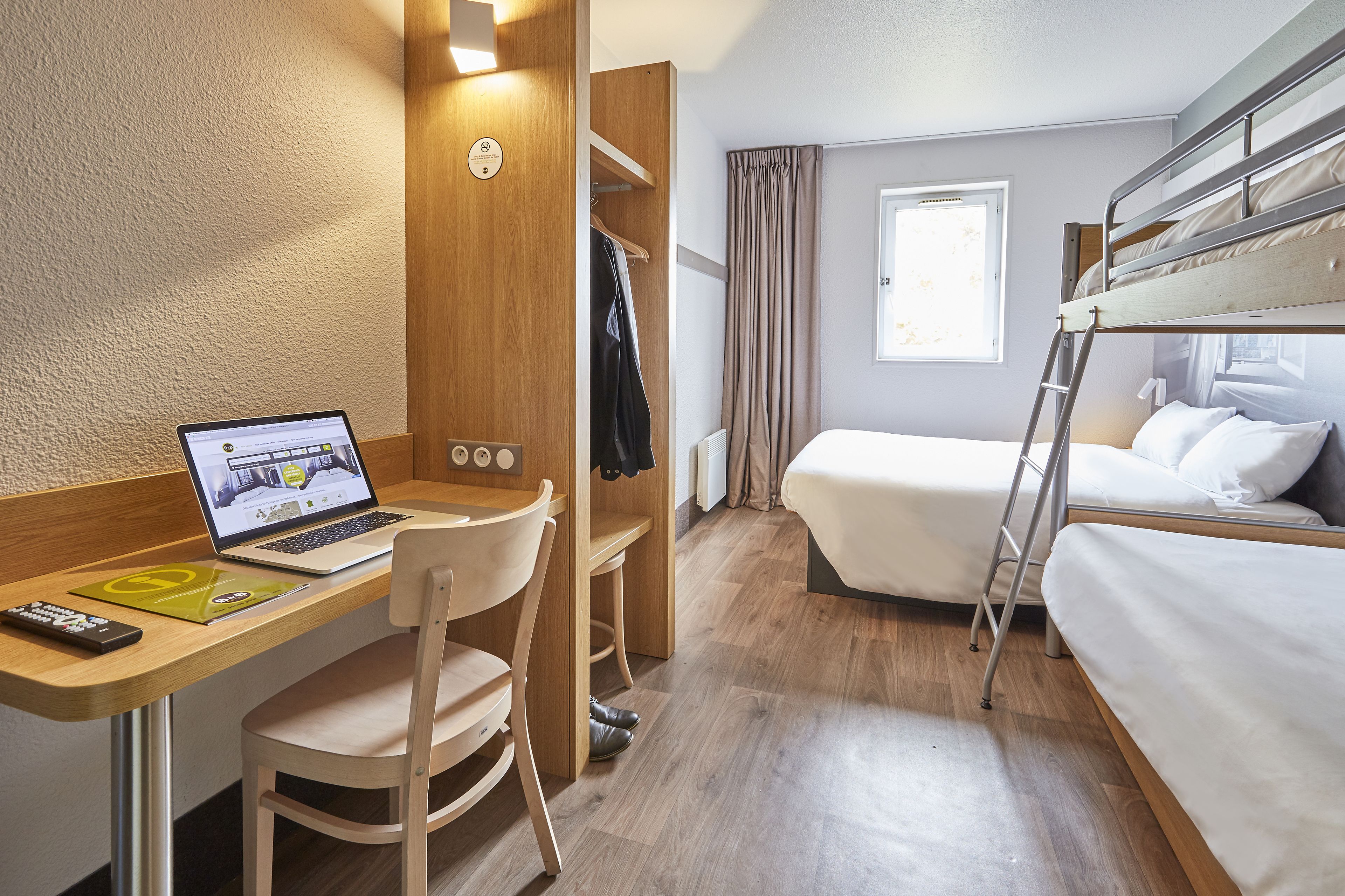 Een compacte hotelkamer met een stapelbed, een bureau met een laptop, een stoel, een kledingkast en een raam met gordijnen.