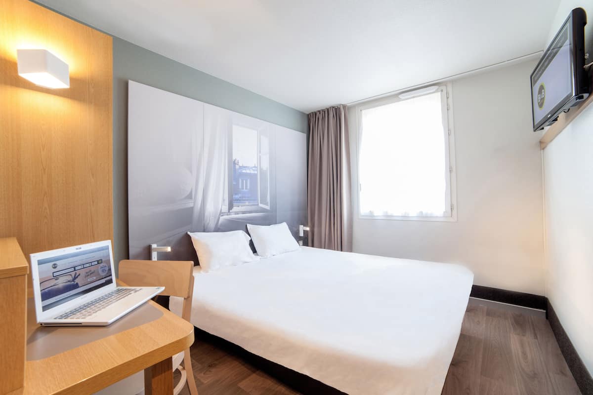 Een hotelkamer met een bed, een bureau met een laptop, een aan de muur bevestigde televisie en een raam met gordijnen.